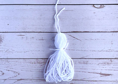Grab your yarn once again to make the head of the ghost.  In a double knot, wrap a piece of yarn that is 7′′ long around the top quarter of the tassel. This creates the ghost's "head." Then, the "neck" will be created by wrapping the yarn's remaining piece around a few times.  After double-knotting the yarn, trim any loose ends.